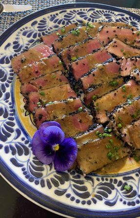 Sesame-crusted seared ahi tuna slices drizzled with savory sauce, sprinkled with black sesame and chives, garnished with a purple edible viola on a blue patterned ceramic plate