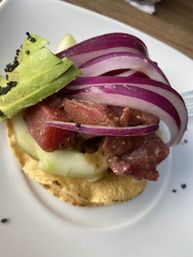Close-up of a colorful tuna tostada: marinated raw tuna tartare on a crispy tortilla topped with cucumber slices, sliced avocado, red onion ribbons and black sesame seeds on a white plate.