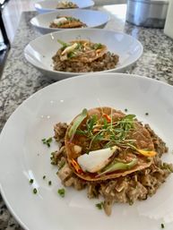 Row of white bowls on a speckled granite counter holding gourmet appetizers — crispy golden rounds topped with microgreens, edible flower petals and a white garnish, set on a creamy diced savory mix.