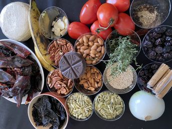 Colorful spread of Mexican mole ingredients on a black counter: dried chilies and chiles, chocolate disk, tomatoes, garlic, white onion, almonds, pecans, sesame and pumpkin seeds, cinnamon sticks, raisins, tortillas and a plantain.