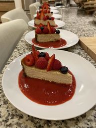 Row of cheesecake slices topped with raspberries, strawberry slices and blueberries, each served on bright strawberry coulis on white plates lined up on a granite kitchen countertop
