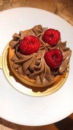 Close-up of a decadent chocolate mousse tart piped in swirls, topped with three fresh raspberries on a gold base, served on a white plate