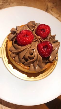 Close-up of a decadent chocolate mousse tart piped in swirls, topped with three fresh raspberries on a gold base, served on a white plate