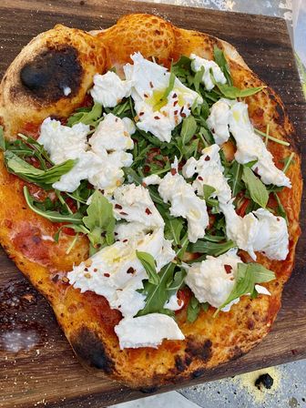 Rustic wood-fired Neapolitan-style pizza with charred golden crust, tomato sauce, peppery arugula, torn burrata, chili flakes and drizzle of olive oil on a wooden board