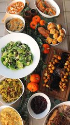 Festive autumn/Thanksgiving dinner spread with sliced roast beef and glazed baby potatoes on a wooden board, green apple salad, cranberry sauce, baked mac and cheese, biscuits, mashed sweet potato, and mini pumpkins as table décor.