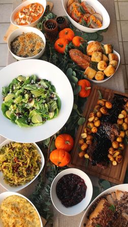 Festive autumn/Thanksgiving dinner spread with sliced roast beef and glazed baby potatoes on a wooden board, green apple salad, cranberry sauce, baked mac and cheese, biscuits, mashed sweet potato, and mini pumpkins as table décor.