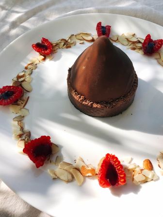 Sunlit chocolate dome tart on a white plate, ringed with halved raspberries filled with jam and scattered sliced almonds — elegant plated dessert.