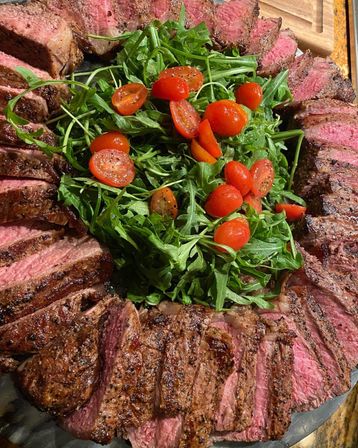 Platter of sliced medium-rare grilled steak arranged in a ring around peppery arugula topped with halved cherry tomatoes and cracked black pepper