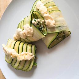 Zucchini ribbon rolls filled with herbed ricotta and fresh herbs, topped with dollops of creamy sauce on a white plate