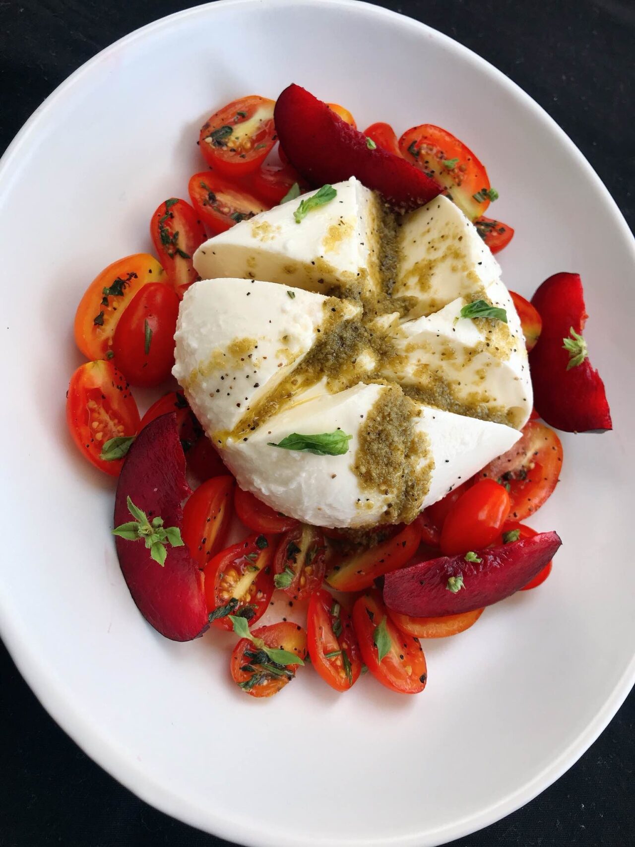Creamy burrata drizzled with pesto over halved cherry tomatoes and plum wedges, garnished with fresh basil on a white plate.