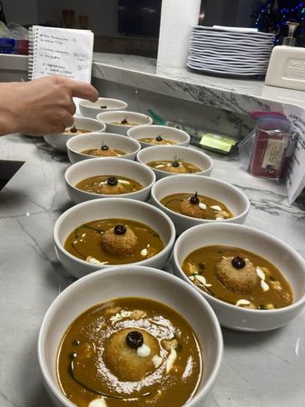 White bowls of golden soup lined up on a marble countertop, each topped with a crispy croquette, cream drizzle and a dark sauce dot — plated at a professional kitchen prep station.
