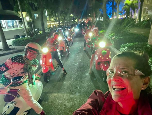Selfie of a smiling rider leading a nighttime festive scooter parade down a palm-lined street, riders on brightly lit red and white scooters wearing holiday sweaters, string lights and Santa hats.