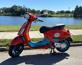 Bright red and teal retro scooter parked on a sunny lakeside road with grassy waterfront and trees in the background