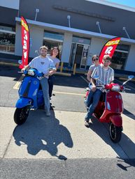Two smiling couples on a blue and a red Vespa scooter parked outside a scooter rental storefront in a sunny parking lot.