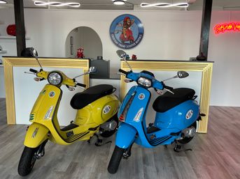 Two retro-style scooters, one yellow and one blue, parked inside a bright St. Petersburg scooter rental shop with a gold-trimmed counter, round logo on the wall, and neon sign