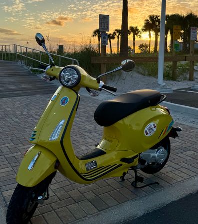 Yellow scooter parked on a brick coastal boardwalk near sand dunes and palm trees, glowing under a golden beach sunset with the ocean in the background