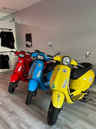 Bright yellow, blue, and red Vespa-style scooters lined up in an indoor showroom with gray walls, wood-look flooring, and a clothing rack in the background.
