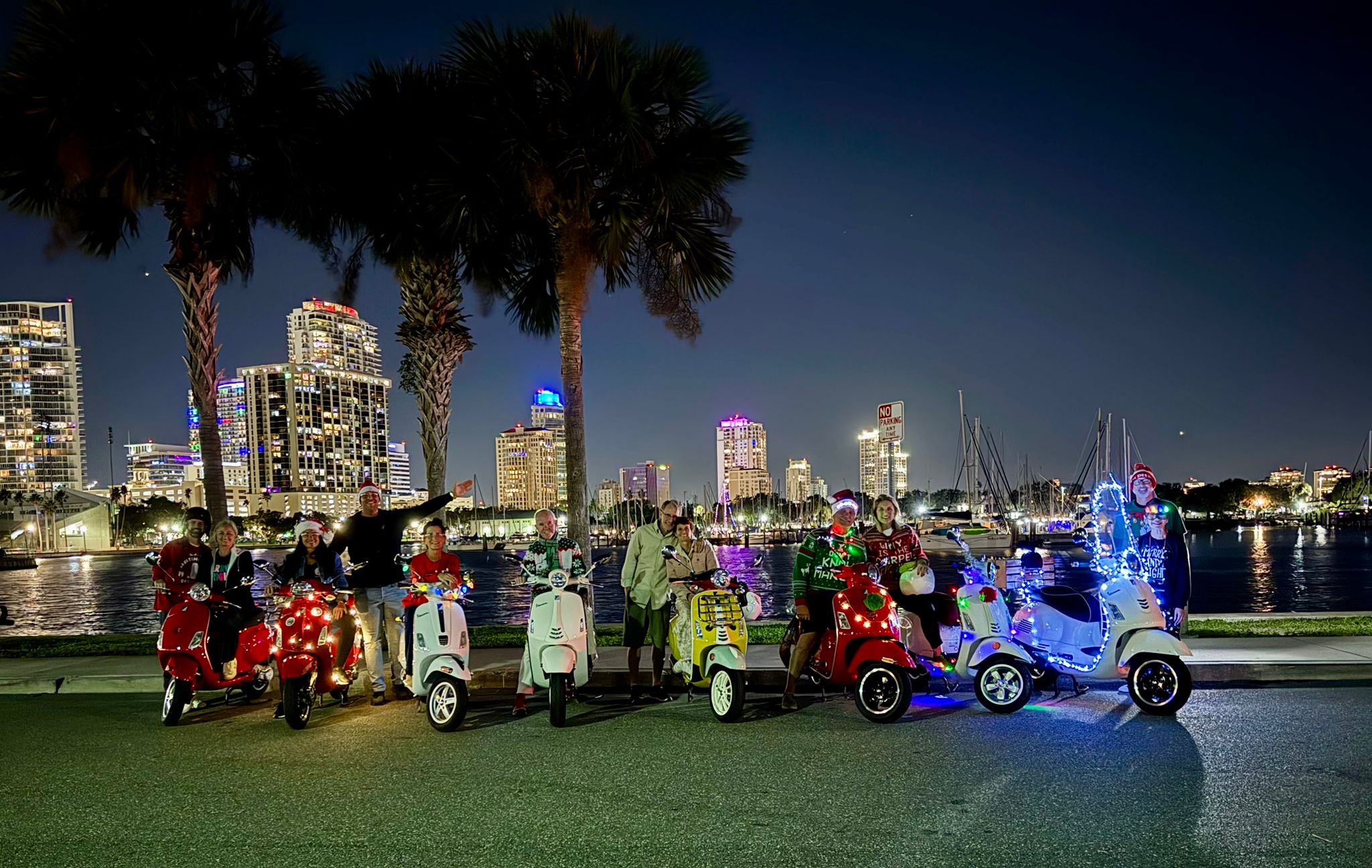 Festive group on decorated motor scooters with holiday lights parked by palm trees along a waterfront marina at night with an illuminated city skyline