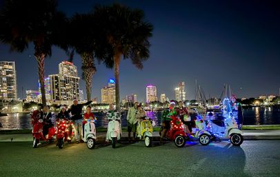 Festive group on decorated motor scooters with holiday lights parked by palm trees along a waterfront marina at night with an illuminated city skyline