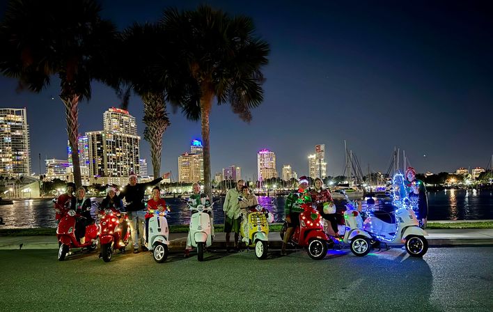 Festive group on decorated motor scooters with holiday lights parked by palm trees along a waterfront marina at night with an illuminated city skyline