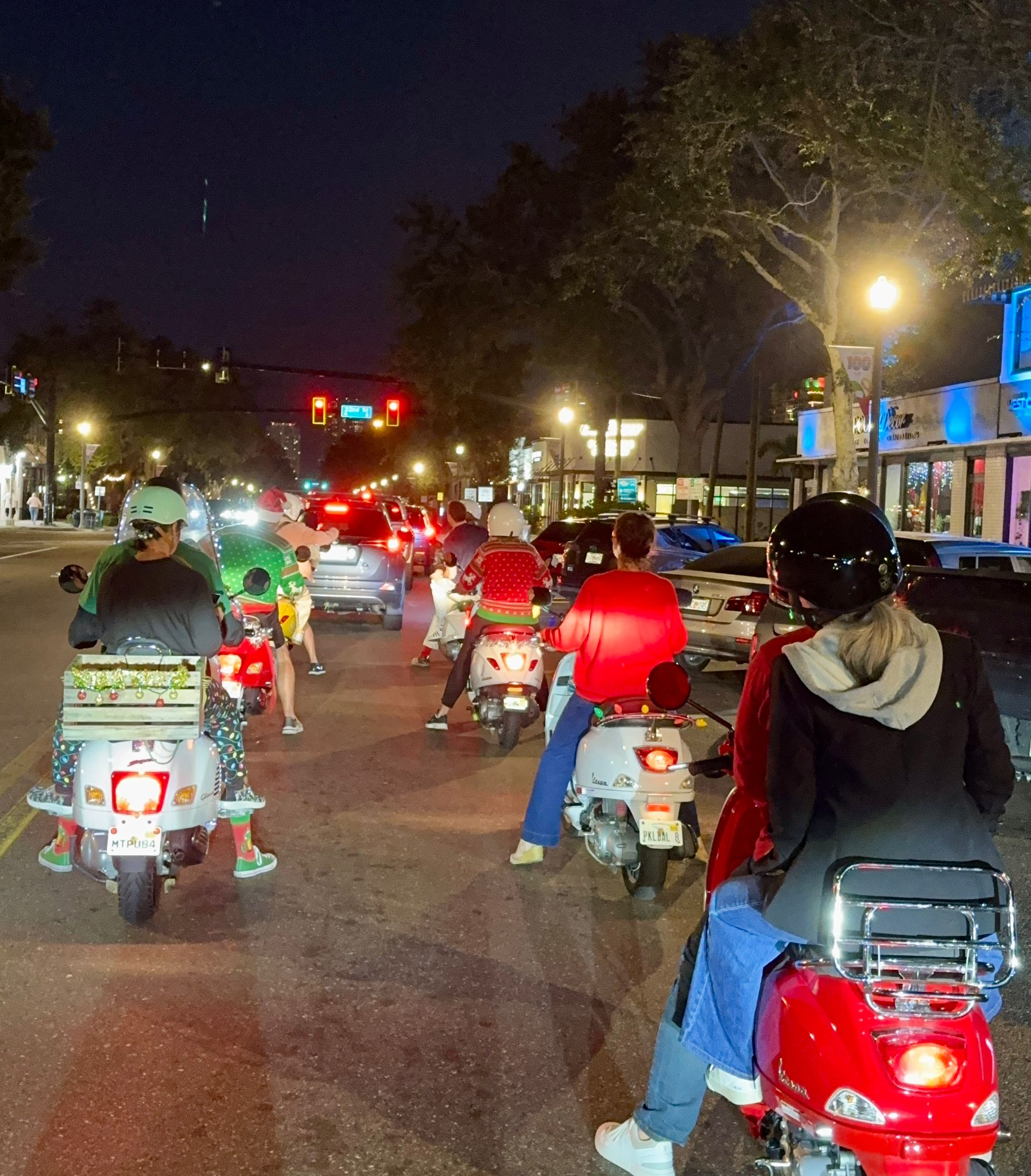 Group of scooter riders in helmets and holiday sweaters stopped at a red light on a downtown street at night, lined with shops and streetlights.