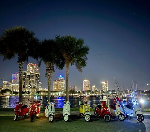 Line of colorful scooters decorated with string lights parked under palm trees along a waterfront promenade at night, with a lit city skyline, marina and sailboats reflecting on the water.