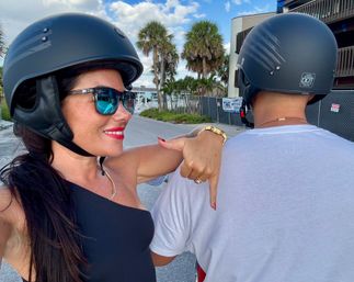 Smiling rider in sunglasses and a black helmet takes a selfie, pointing at a fellow helmeted rider on a scooter along a palm-lined coastal street under a bright blue sky.