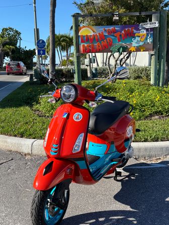 Red and turquoise scooter parked at a sunny coastal entrance with palm trees and a colorful "Livin' on Island Time" Sunset Beach sign