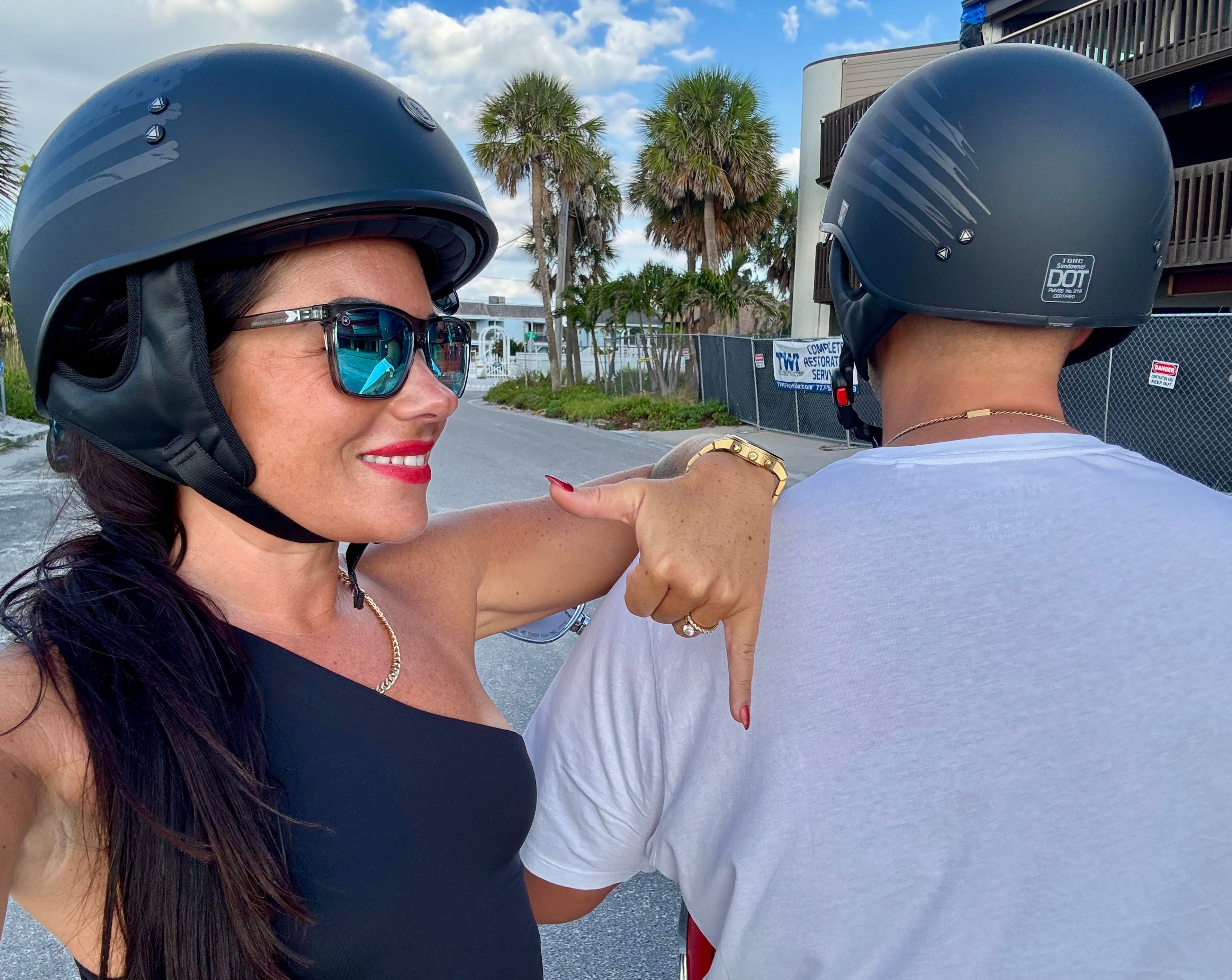Smiling rider in sunglasses and a black helmet takes a selfie, pointing at a fellow helmeted rider on a scooter along a palm-lined coastal street under a bright blue sky.