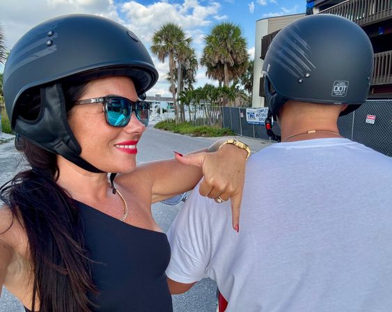 Smiling rider in sunglasses and a black helmet takes a selfie, pointing at a fellow helmeted rider on a scooter along a palm-lined coastal street under a bright blue sky.