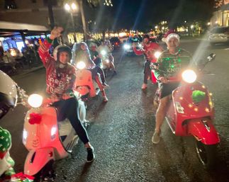 Group of riders on decorated scooters on a downtown street at night wearing holiday sweaters, Santa hats and string lights.