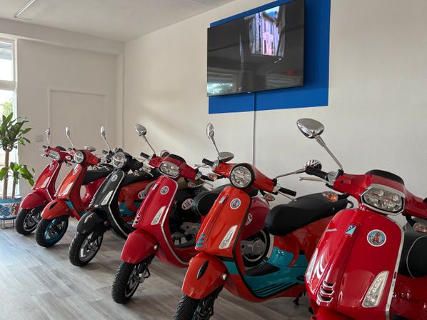 Row of bright red retro-style scooters (one black) lined up indoors on light wood floors in a sunlit showroom with a potted plant and wall-mounted TV