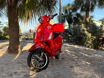 Bright red scooter parked on sandy coastal path beneath palm trees, sunlit tropical shrubs and beach houses in the background