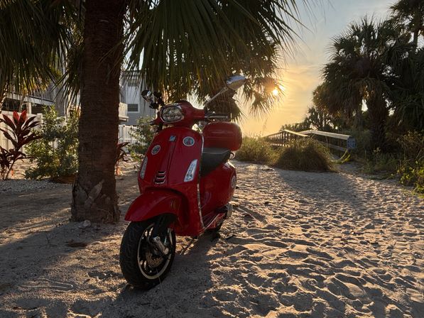 Red scooter chilling on a sandy beach under palm trees at golden sunset, with footprints in the sand and a wooden boardwalk nearby