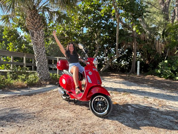 Happy rider on a red scooter parked on a sandy coastal path beneath palm trees, waving and smiling on a sunny tropical beachside day.