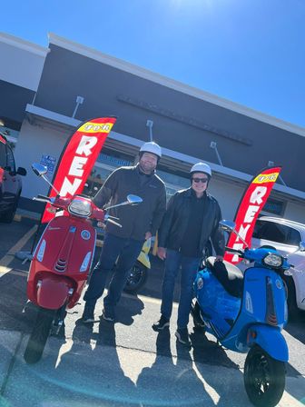 Two people wearing helmets stand with red and blue Vespa-style scooters in front of a scooter rental storefront with red 'RENT' flags on a bright sunny day.