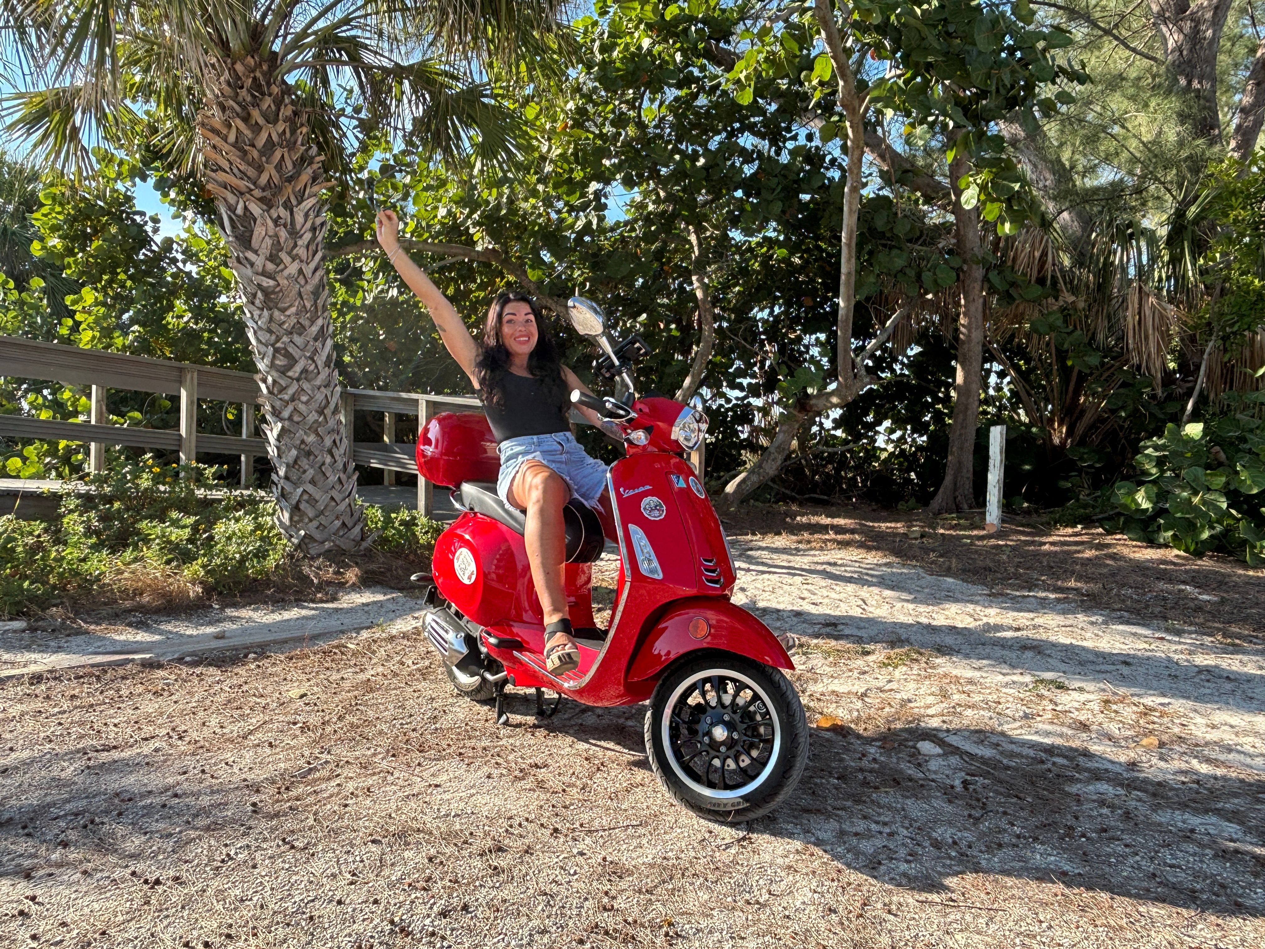 Cheerful rider on a bright red Vespa-style scooter waving beside palm trees and a sandy coastal boardwalk on a sunny tropical day.
