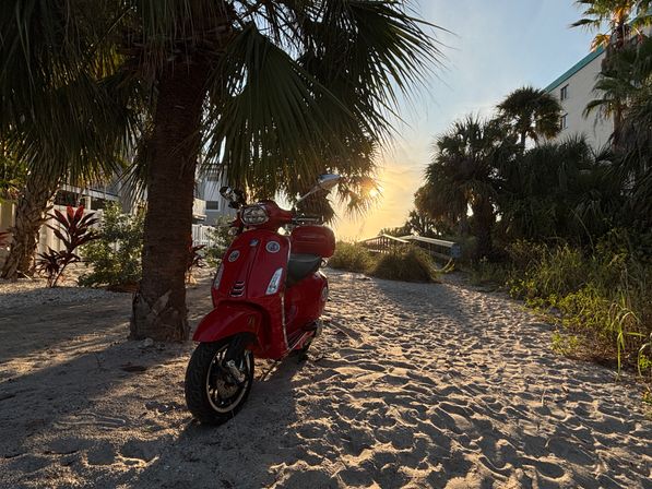 Red scooter parked on a sandy beach path under palm trees at golden sunset, coastal boardwalk and tropical foliage in the background.