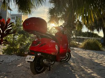 Sun-kissed red scooter parked on a sandy coastal path under palm trees at golden sunset, tropical foliage and a boardwalk in the background.
