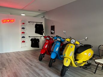 Three retro-style scooters—yellow, blue, and red—lined up in a bright modern showroom with wood-look flooring, a white display wall of hats and hanging black t-shirts, and a pink neon wall sign.