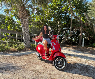 Joyful rider on a shiny red scooter parked on a sandy beach path beneath palm trees and lush coastal greenery, sunny tropical vibe.