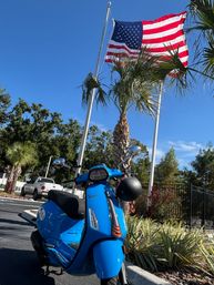 Bright blue Vespa scooter parked in a sunny lot with a black helmet on the handlebar, waving American flag overhead and palm trees against a clear blue sky