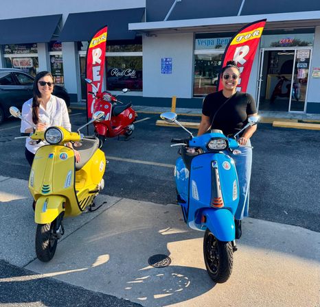 Two smiling women wearing sunglasses pose on bright yellow and blue retro scooters parked outside a scooter rental storefront on a sunny day.