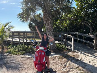 Smiling person on a red scooter at a sunny tropical beach boardwalk beneath a palm tree, sandy foreground and distant ocean horizon