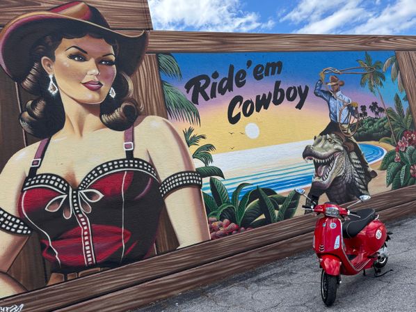 Red scooter parked in front of a vibrant street-art mural of a pinup cowgirl and a cowboy lassoing a dinosaur on a tropical beach with the phrase 'Ride 'em Cowboy'.