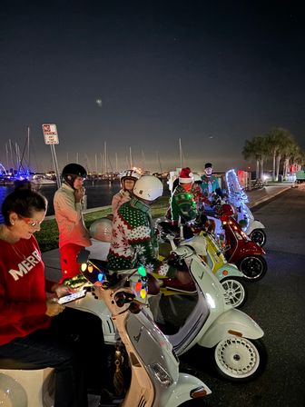Group of people on festively decorated scooters at a waterfront marina at night, wearing Christmas sweaters and Santa hats with sailboats and palm trees in the background.