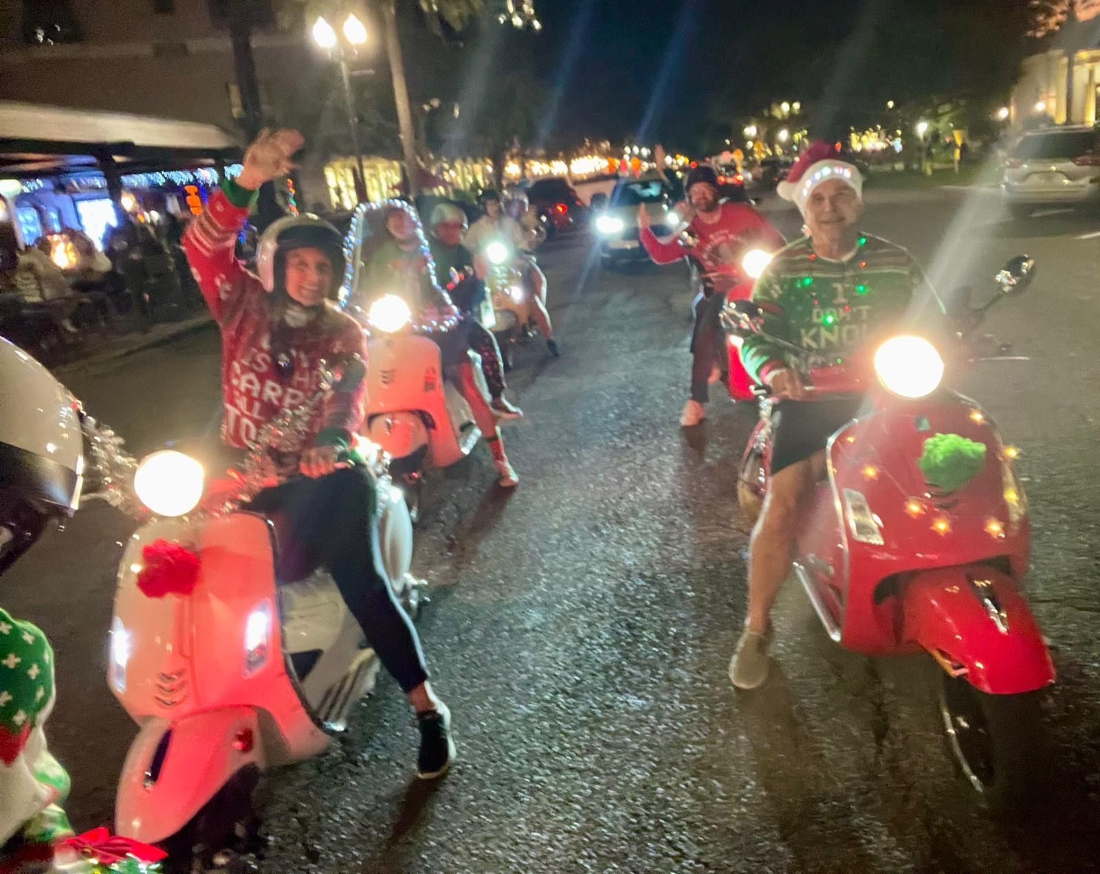 Festive holiday scooter ride on a downtown street at night — riders on decorated Vespas wearing Santa hats and Christmas sweaters, waving under glowing storefront lights
