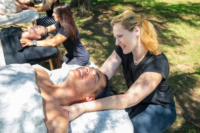 Smiling client receives a neck and shoulder massage from a therapist on a portable table at an outdoor park wellness event with dappled sunlight on the grass.