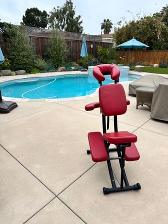 Red portable massage chair on a concrete poolside patio beside a curved backyard swimming pool with blue umbrellas, covered lounge furniture, green shrubs and a wooden fence.
