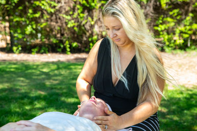 Blonde woman giving a gentle head-and-neck massage to a person lying on a blanket in a sunlit park with green trees